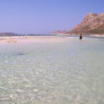 Person walking in shallow, clear waters with mountains in the background.
