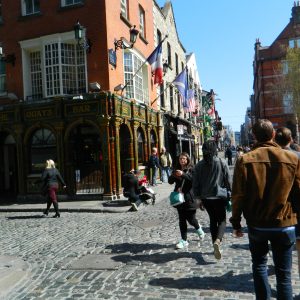 People walking on a cobblestone street lined with colorful buildings and flags.